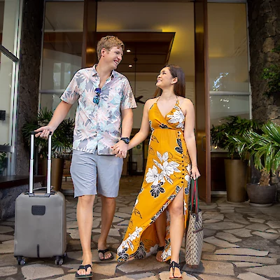 A couple is walking outside a building, holding hands and a suitcase, dressed in summer attire and smiling at each other.