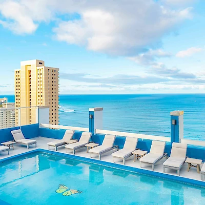 Rooftop pool with lounge chairs, overlooking ocean and tall buildings under a partly cloudy sky.