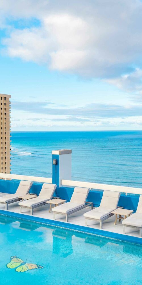 Rooftop pool with lounge chairs, overlooking ocean and tall buildings under a partly cloudy sky.