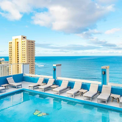A rooftop pool with lounge chairs overlooks a cityscape and ocean under a partly cloudy sky.