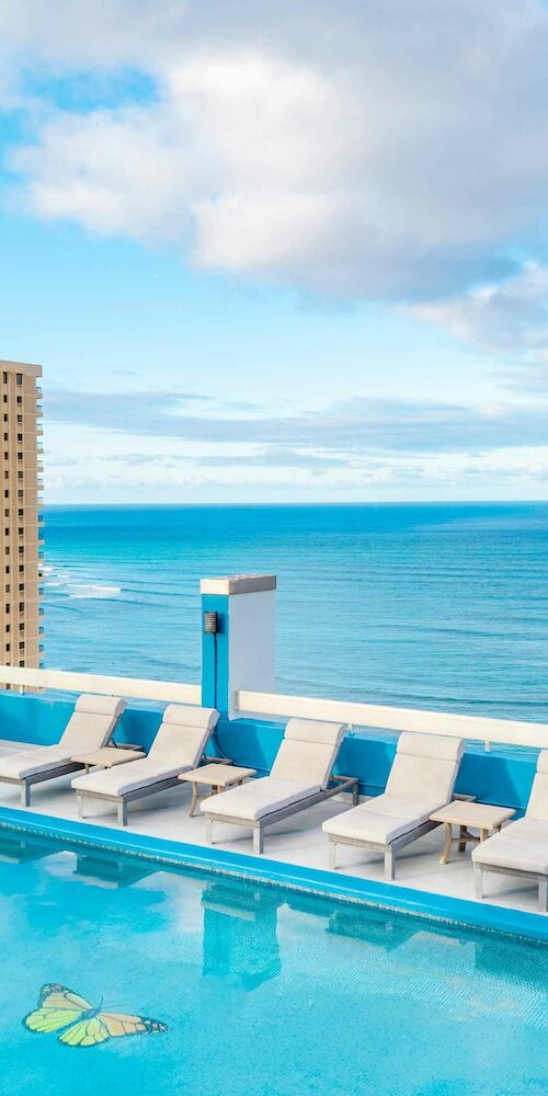 A rooftop pool with lounge chairs overlooks a cityscape and ocean under a partly cloudy sky.