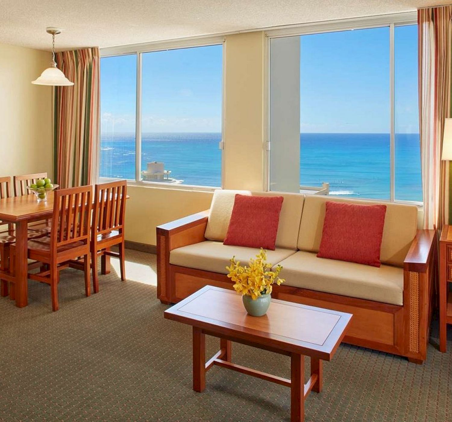 A cozy, sunlit living room with a wooden dining set, sofa, coffee table, and ocean view. Decor includes striped curtains and a wall art piece.