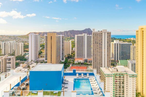 The image shows a cityscape with high-rise buildings, a rooftop pool, and a distant mountain under a clear blue sky.