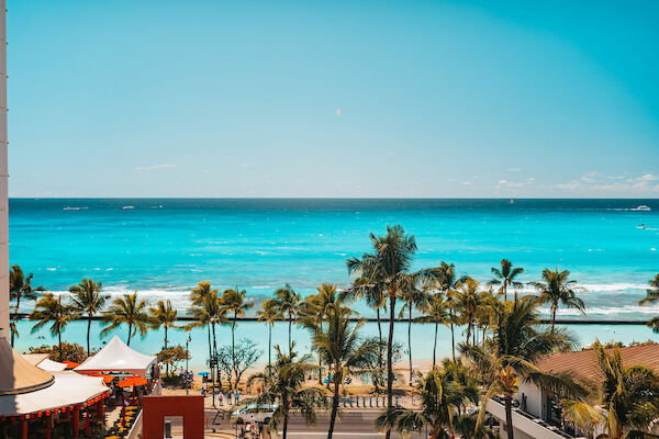 A scenic beach view with turquoise water, palm trees, and a few buildings in the foreground on a bright, sunny day, ending the sentence.