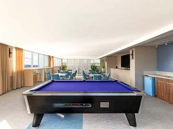 A recreation room with a blue pool table, chairs, tables, a TV, and a kitchenette. Large windows bring in natural light.