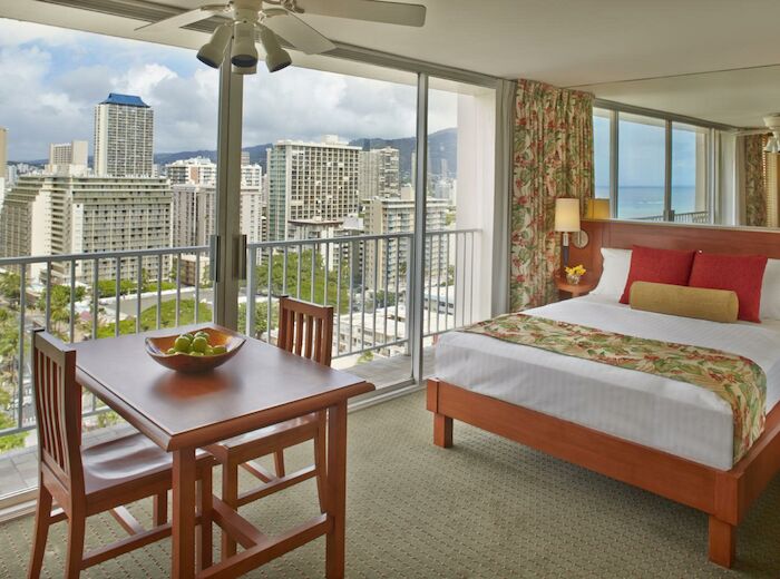A cozy hotel room with a bed, dining table, and city view through large windows. The room has a ceiling fan and a modern decor.