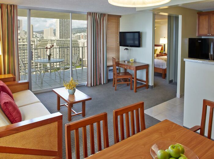 The image shows a bright and modern living area with a sofa, dining table, and balcony view. The space includes a kitchenette and a wall-mounted TV.
