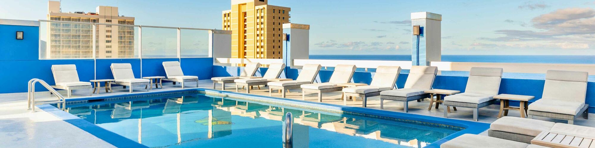 Rooftop pool with lounge chairs and a clear blue sky, surrounded by buildings in the background, under a few clouds.