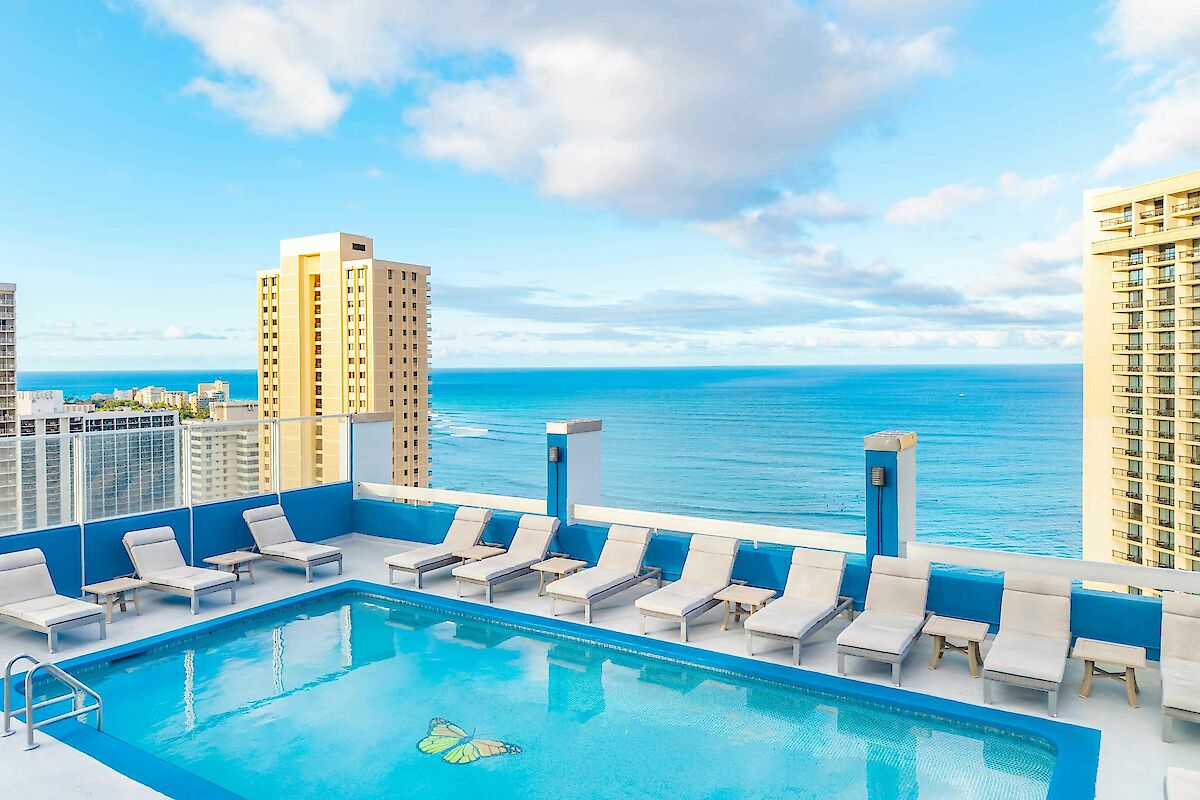 A rooftop pool with lounge chairs overlooks a scenic ocean view, surrounded by tall buildings under a partly cloudy sky.