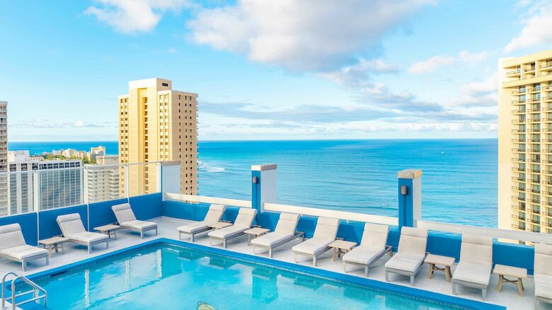 A rooftop pool with lounge chairs overlooks a scenic ocean view, surrounded by tall buildings under a partly cloudy sky.
