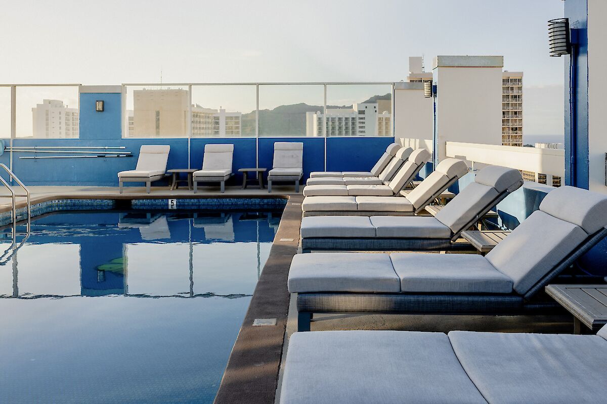 A rooftop swimming pool with multiple lounge chairs lined up next to it. A cityscape is visible in the background under a clear sky.