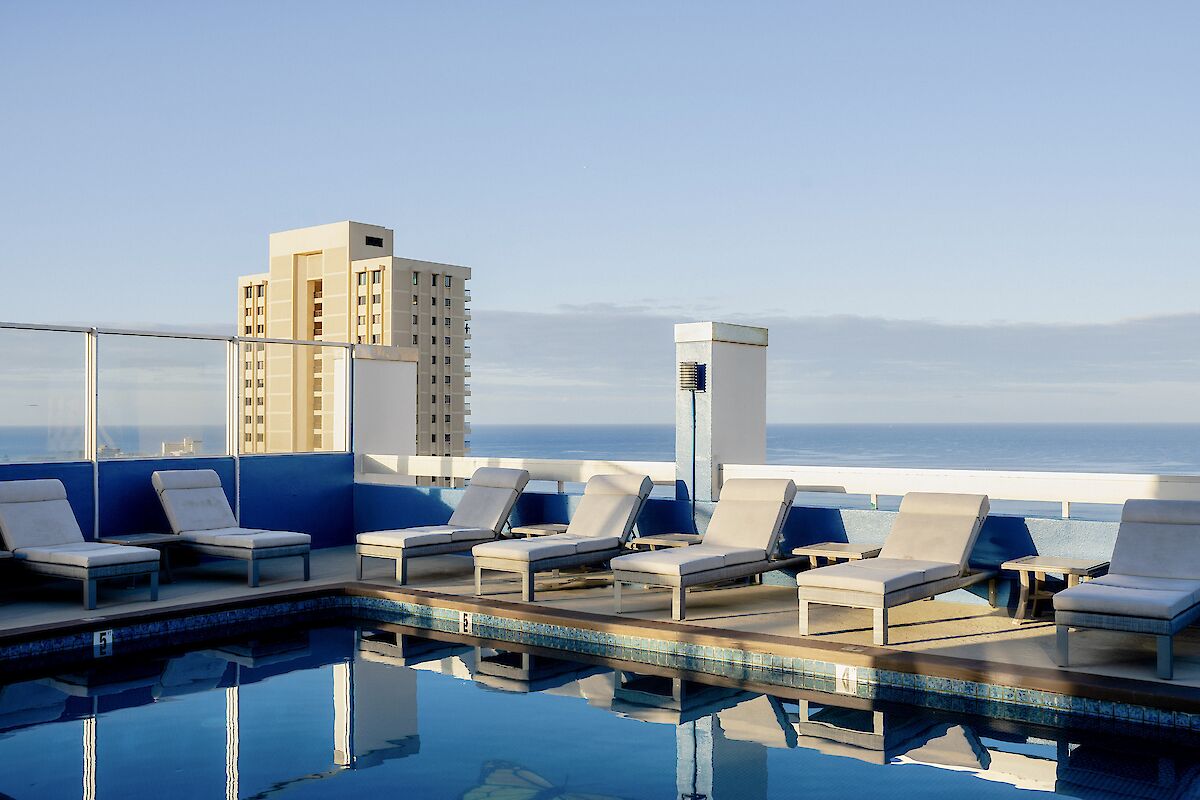 The image shows a rooftop pool with several lounge chairs around it, overlooking a cityscape and the ocean under a clear blue sky.