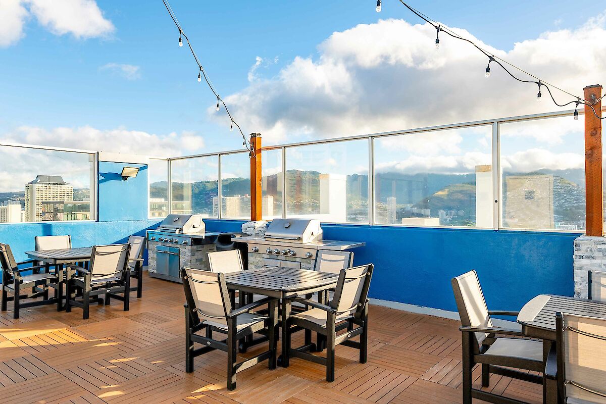 A rooftop patio with tables, chairs, BBQ grills, string lights, and scenic views under a blue sky with some clouds.