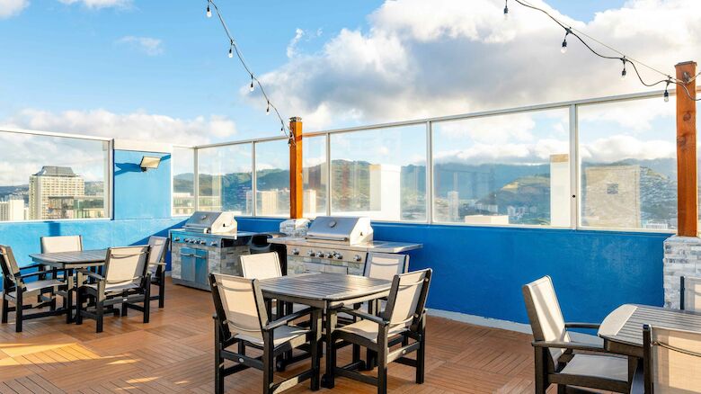 A rooftop patio with tables, chairs, BBQ grills, string lights, and scenic views under a blue sky with some clouds.