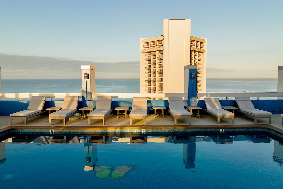A rooftop pool with lounge chairs beside it overlooks a tall building and the ocean in the distance.