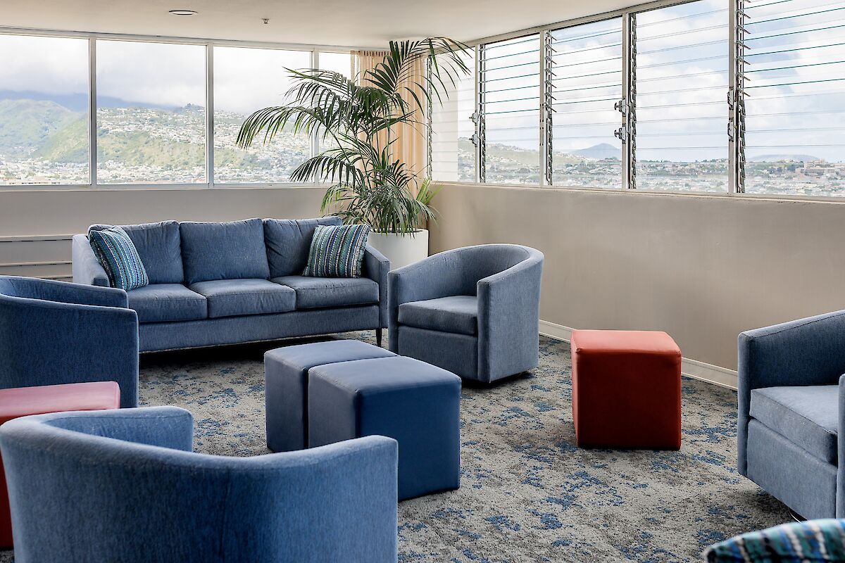 The image shows a sitting area with a blue sofa, blue chairs, and colorful ottomans near large windows with a scenic view outside.
