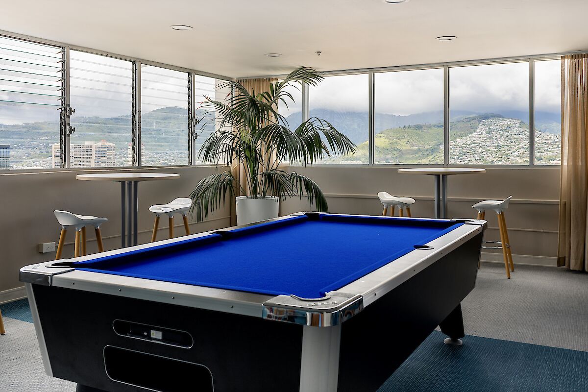 The image shows a room with a pool table, bar stools around high tables, a potted palm, and large windows with mountain views outside.