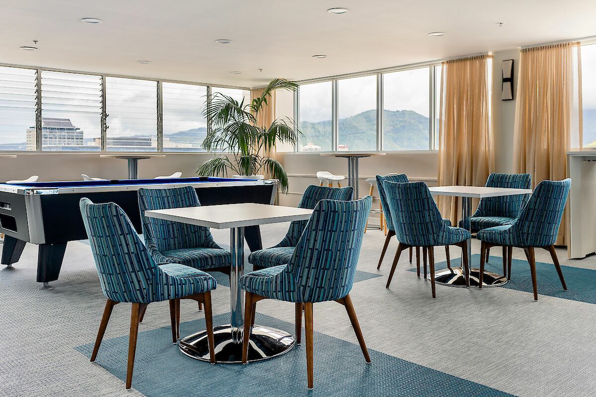 A modern lounge area with tables and blue chairs, a pool table, large windows with view of mountains, and a potted plant in the corner.