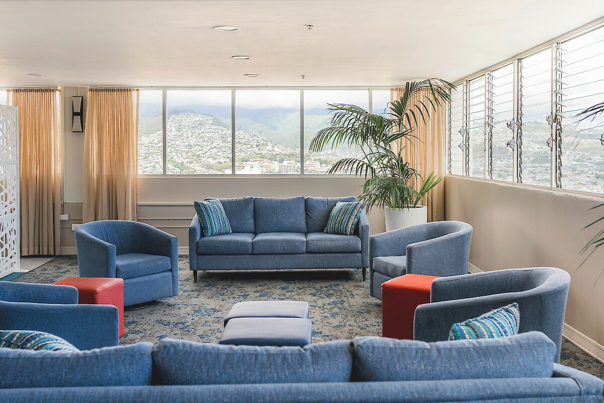 A spacious living room with blue sofas, two red ottomans, large windows, potted plants, and a view of mountains in the distance.