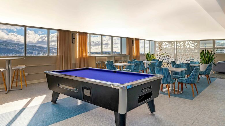 A modern room with a pool table, chairs, tables, and large windows offering mountain views under a bright ceiling.