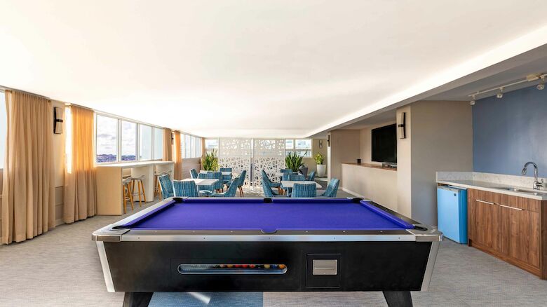 A room with a pool table, seating area, TV, and kitchenette. Bright decor with blue accents and natural light enhance the space.