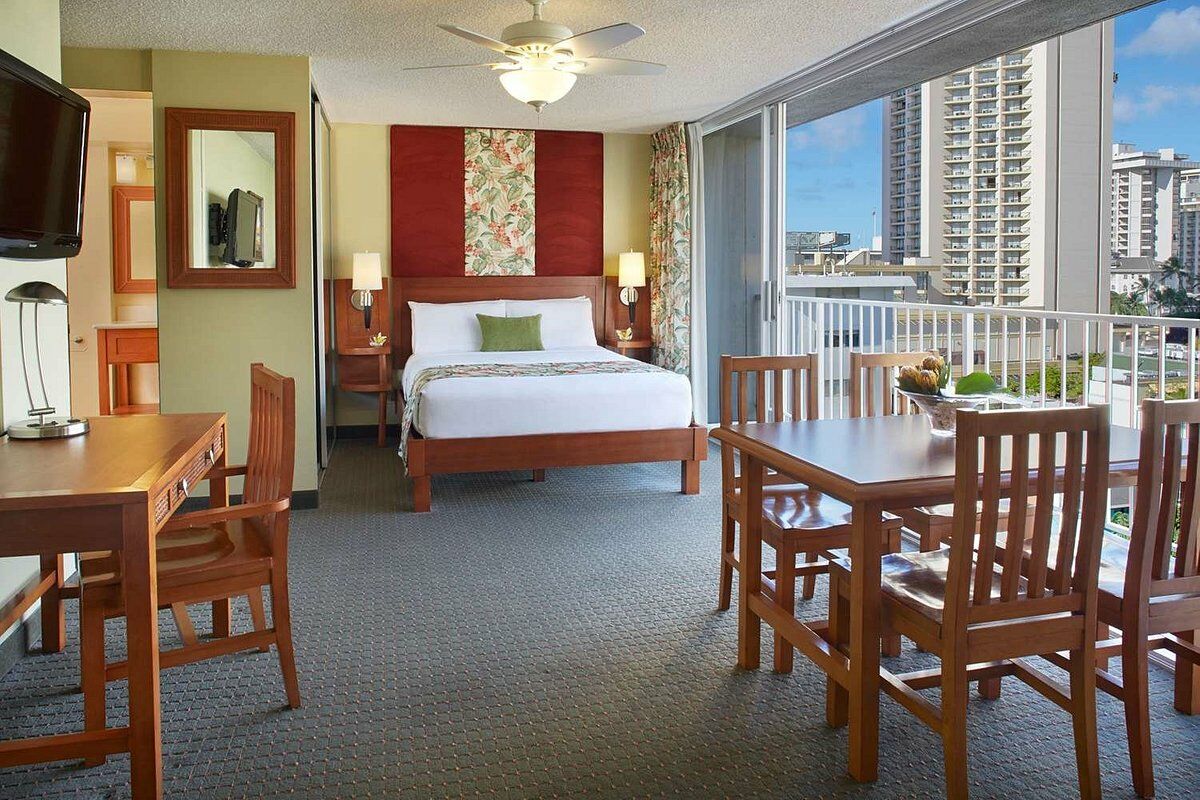 The image shows a well-furnished hotel room with a bed, dining table, chairs, desk, TV, ceiling fan, and a balcony with a view of buildings and a blue sky.
