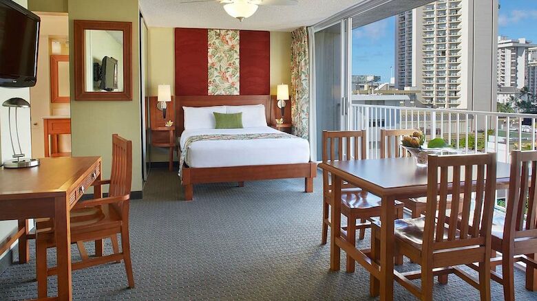The image shows a well-furnished hotel room with a bed, dining table, chairs, desk, TV, ceiling fan, and a balcony with a view of buildings and a blue sky.