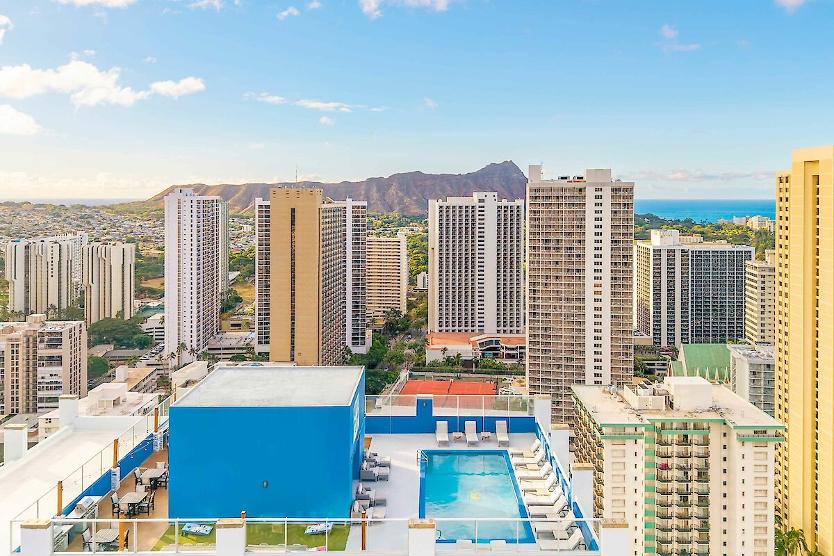 A cityscape with tall buildings, a pool on a rooftop, and mountains in the background under a clear blue sky.