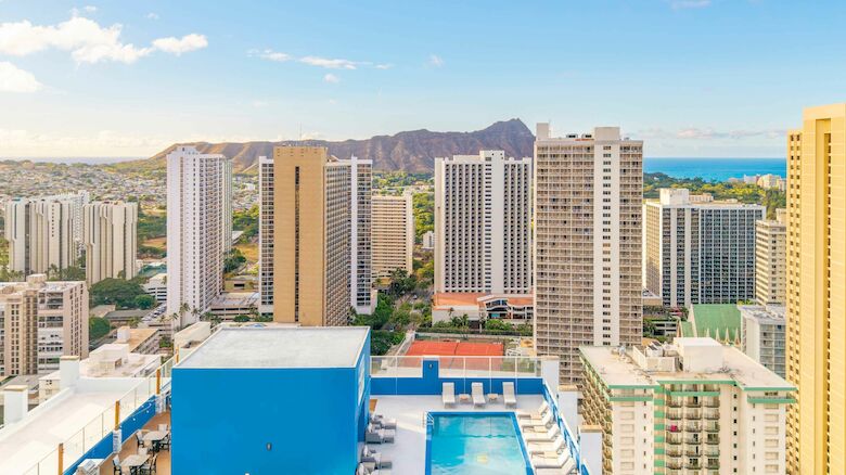 A cityscape with tall buildings, a pool on a rooftop, and mountains in the background under a clear blue sky.