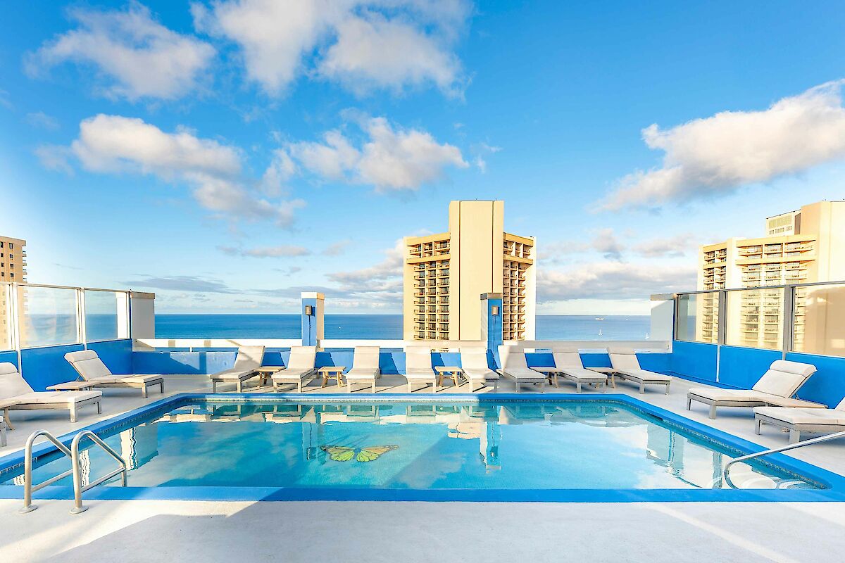 A rooftop pool with lounge chairs overlooks a city skyline and the ocean under a partially cloudy sky.