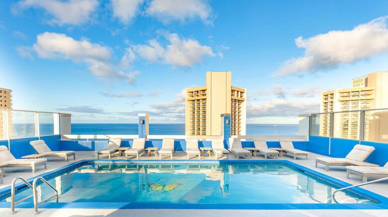 A rooftop pool with lounge chairs overlooks a city skyline and the ocean under a partially cloudy sky.
