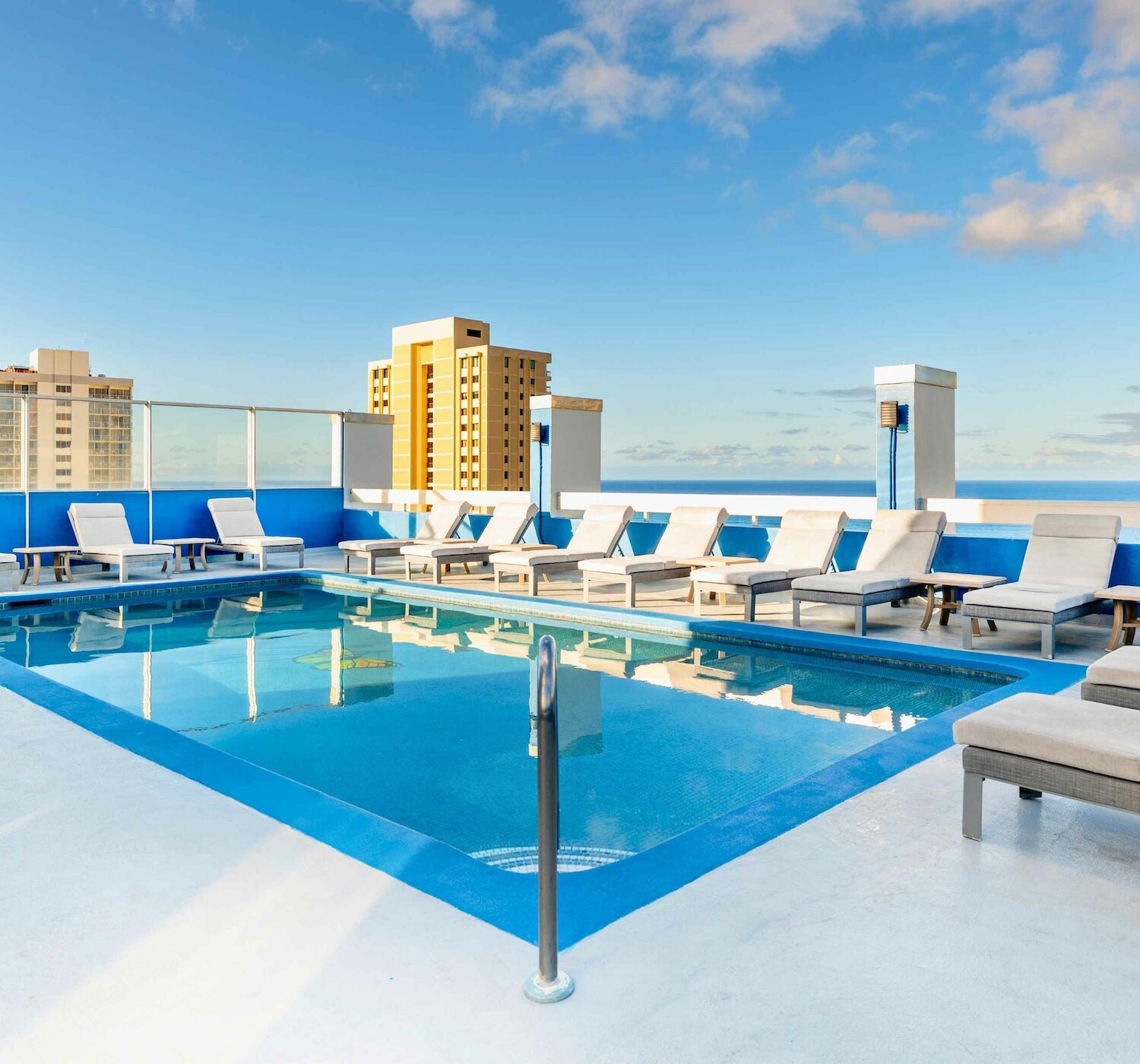 A rooftop swimming pool with lounge chairs, surrounded by a blue sky and city buildings in the background.