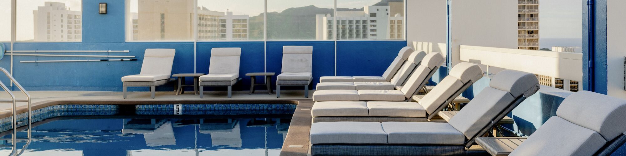 This image depicts a rooftop swimming pool area with several lounge chairs and city buildings in the background.