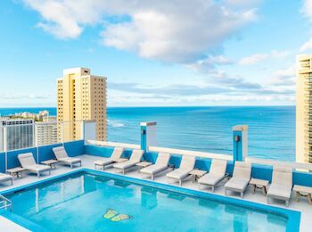 A rooftop pool with lounge chairs overlooks a city skyline and the ocean on a bright, partly cloudy day.