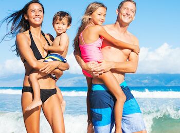 A smiling family of four, in swimwear, is standing at the beach with the ocean waves in the background and clear blue skies overhead.