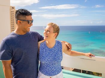 A couple is smiling while posing on a balcony with a scenic ocean view in the background. It appears to be a sunny day.