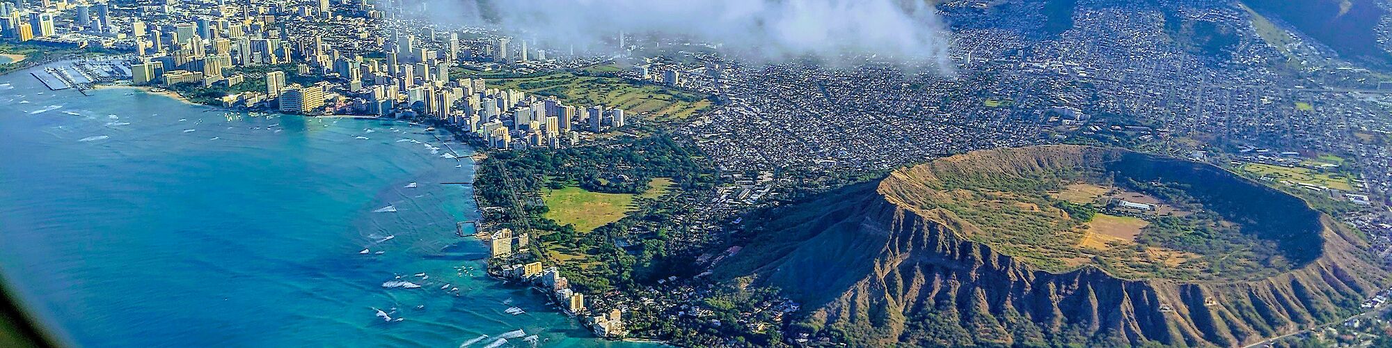 Aerial view of a coastline with a city, a volcanic crater, and ocean, taken from an airplane window with a wing visible.