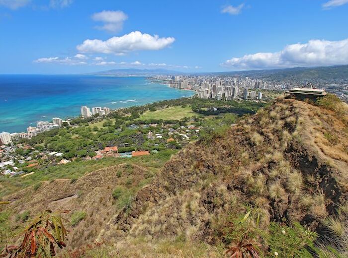 The image shows a coastal city with a sprawling urban area next to the ocean, surrounded by lush greenery and mountains, under a clear blue sky.