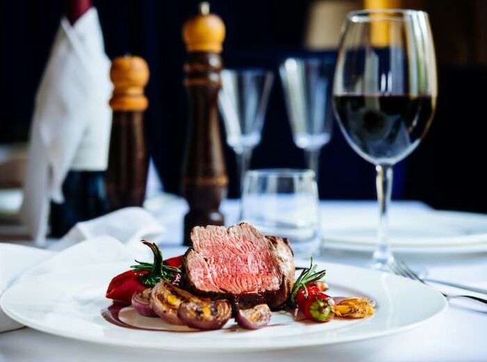 The image shows a gourmet meal featuring a medium-rare beef steak, accompanied by garnishments and vegetables, with a glass of red wine on the side.