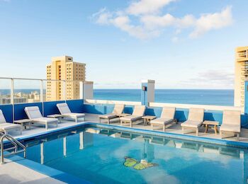 A rooftop pool with lounge chairs and ocean view, set against a backdrop of tall buildings under a partly cloudy sky.