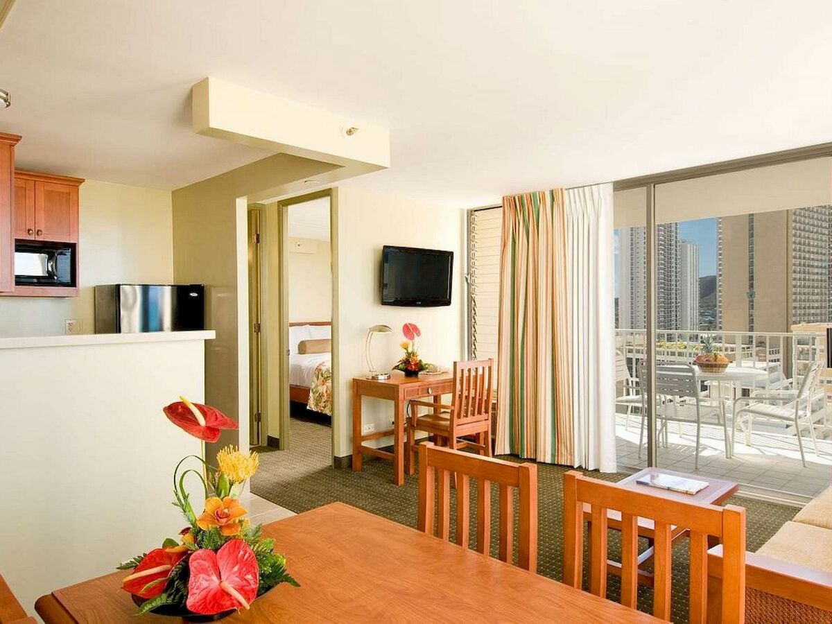 This image shows a modern hotel room with a seating area, kitchenette, TV, and a balcony with a city view. Decor includes flowers and wooden furniture.