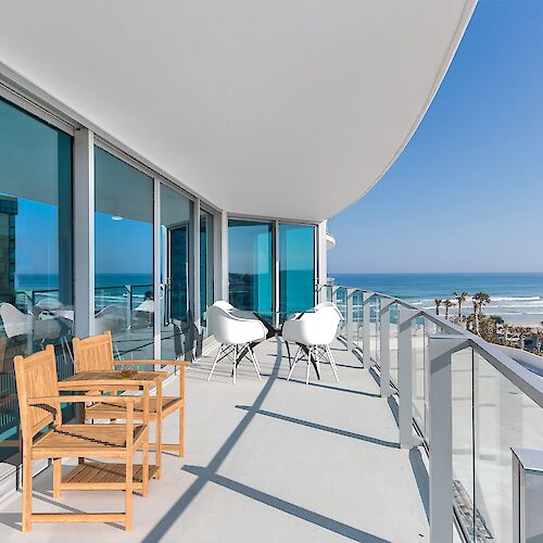 A coastal balcony with wooden and white chairs, glass railing, ocean view, and a modern building nearby on a sunny day.