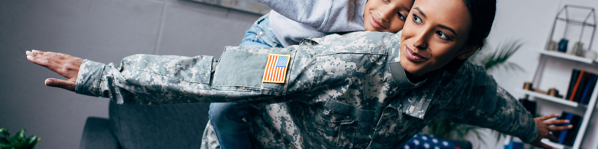 A person in a military uniform holds a child on their back, both with arms outstretched in a playful airplane pose.