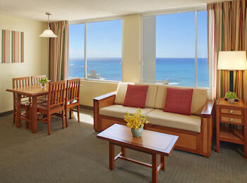A cozy living area with a dining table, a couch, a coffee table, and a great view of the ocean through large windows, decorated in warm tones.