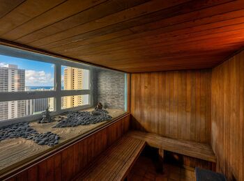 A wooden sauna room with a bench, a large window showing city buildings, and a decorative rock and sand feature.