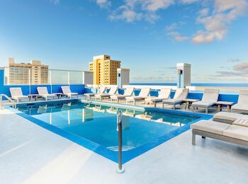 A rooftop pool surrounded by lounge chairs against a backdrop of city buildings and a clear blue sky.