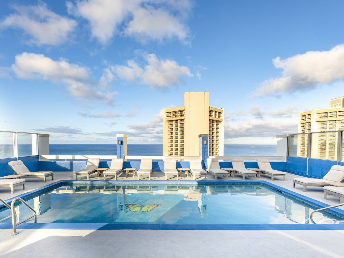 A rooftop pool with lounge chairs, surrounded by buildings, under a blue sky with scattered clouds.