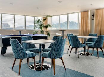 The image shows a modern lounge area with multiple blue chairs, round tables, and a pool table near large windows offering a scenic view.