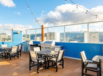 Rooftop patio with tables, chairs, string lights, and grills against a blue wall, with scenic views of the city and mountains in the background.