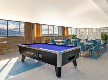 The image shows a lounge with a pool table, blue chairs, tables, and large windows offering a scenic view.